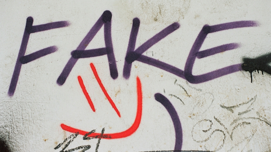 Graffiti auf einer dreckige weißen Häußerwand. „Fake“ steht in Großbuchstaben in dunkellila Farbe auf der Wand, darunter ein roter Smiley.