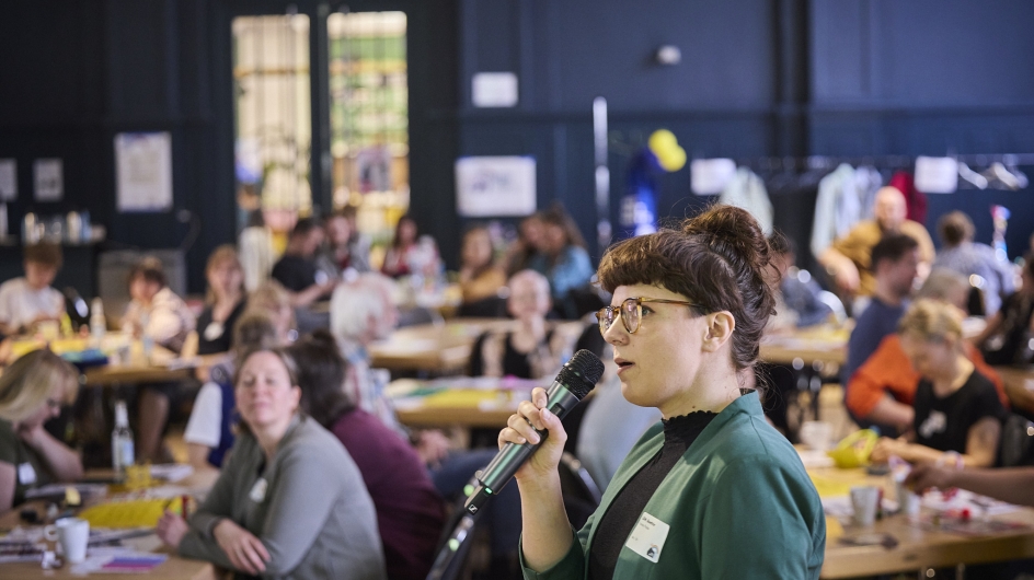 Lilith Boettcher spricht in ein Mikrofon, im Hintergrund sitzen viele Menschen im Publikum 