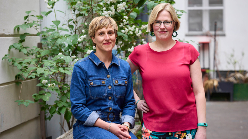 Zwei Personen stehen in einem begrünten Innenhof vor einem blühenden Strauch mit weißen Blüten. Eine sitzt seitlich auf einer niedrigen Mauer und trägt ein blaues Jeanskleid, die andere steht daneben mit einem roten Oberteil und gemusterter Hose, eine Hand locker an die Hüfte gelegt. Beide blicken in die Kamera und wirken ruhig und freundlich. Im Hintergrund sind helle Hauswände und Fenster zu sehen, die dem Bild eine offene, urbane Atmosphäre geben.