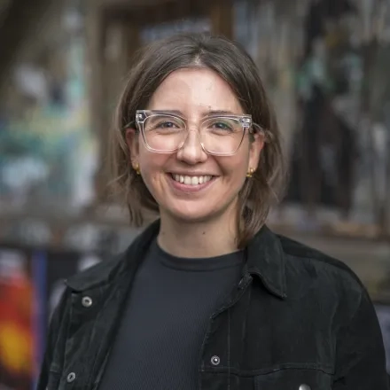 Elisabeth Rieber trägt eine Brille und die Haare nach hinten gebunden in einem schwarzen Hemd vor einem Backsteingebäude mit Graffiti. 