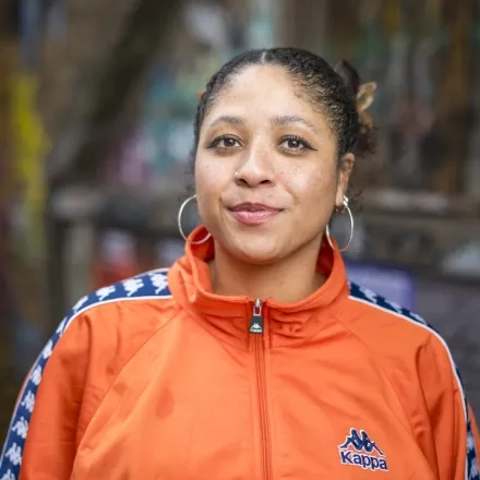 Peace Solomon Obong steht in einer orangefarbenen Jacke vor einem Backstein Gebäude mit Graffiti und schmunzelt leicht in die Kamera. Ihre Haare sind nach Hinden gebunden.