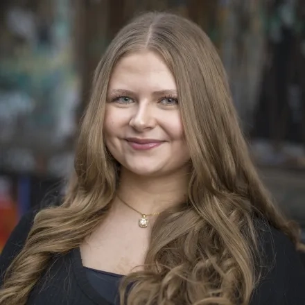 Sabrin Fouani steht vor einer mit Graffiti besprühter Backsteinwand. Sie hat dunkelblonde lange Haare die über ihre Schultern fallen und lächelt in die Kamera.